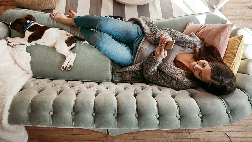 A woman laying on the couch looking at her phone with her dog at her feet
